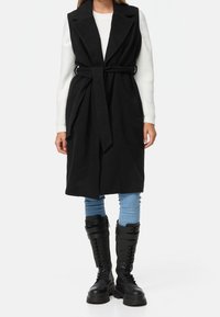 Manteau noir sans manches avec une ceinture, larges revers, texture douce, porté sur un pull blanc, associé à un jean bleu clair et des bottes de combat noires.
