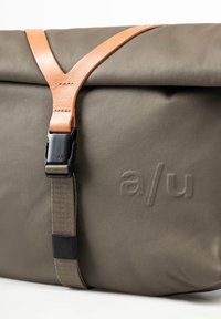 Borsa verde oliva con design pieghevole, dotata di una tracolla in pelle beige e una fibbia nera. Superficie testurizzata con le iniziali del marchio in rilievo "a/u."