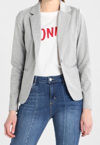Donna che indossa un blazer grigio chiaro sopra una maglietta bianca con testo rosso e jeans in denim blu a vita alta, in piedi con le braccia rilassate.
