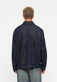 Man met kort krullend haar, gekleed in een donkerblauwe spijkerjack en lichtblauwe jeans, staand tegen een effen witte achtergrond.