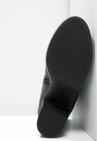 Suola nera di scarpa con superficie texture, caratterizzata da una punta rotonda e tacco basso a blocco. Dettaglio con zip laterale su materiale superiore liscio.