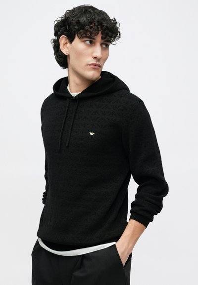 Sweatshirt preta com capuz feita de tecido texturizado, apresentando um padrão subtil, cordão e um pequeno logótipo dourado no peito.