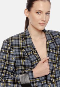 Młoda kobieta z włosami zebranymi w kok, nosi szary, kratkowany żakiet z żółtym haftem nuty muzycznej, trzymając klapę prawą ręką.