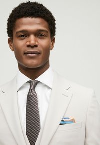 Blazer beige claro sobre una camisa blanca, acompañado de una corbata texturada marrón y blanca. El pañuelo de bolsillo presenta patrones en azul y naranja.