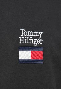 Μαύρο ύφασμα με λευκή κεντητή επιγραφή "Tommy Hilfiger" πάνω από οριζόντια τρίχρωμη λογότυπο: ριγέ σε ναυτικό μπλε, λευκό και κόκκινο.