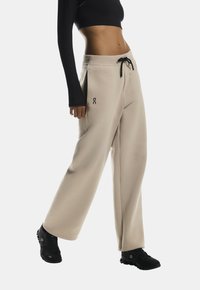 Beige brede sweatpants med snor i taljen, sidelommer og en sort logo detalje. Stoffet ser blødt og tekstureret ud.