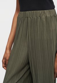 Nahaufnahme einer Person, die hoch taillierte, olivgrüne, plissierte Hosen mit weitem Bein und ein schwarzes ärmelloses Oberteil trägt.