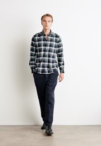 Fiatal férfi sétál bent, fekete-fehér kockás ingben, sötét nadrágban és fekete bőrcipőben, egy sima fehér fal előtt.