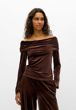 Un mannequin pose sur un fond blanc, portant un haut en velours marron foncé à épaules dénudées et un pantalon assorti.