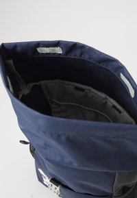 Marinblå tygryggsäck med rullöppning, grått innerfoder, sidoficka och justerbara detaljer.