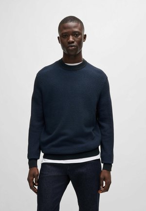 Maglione a trama blu navy con colletto girocollo a coste, maniche lunghe e polsini neri a contrasto. Indossato sopra una camicia bianca a strati.