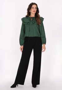 Grüne Bluse mit einem Rüschensaum und Krawattendetails, langen Ärmeln mit elastischen Bündchen, kombiniert mit schwarzen weiten Hosen und Absatzschuhen.