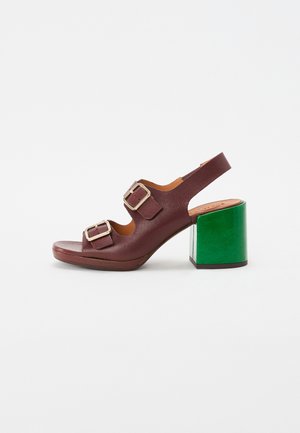 Sandal in pelle marrone con due fibbie argentate, design aperto sulla punta e tacco verde a blocco. Presenta una texture liscia e uno stile minimalista.