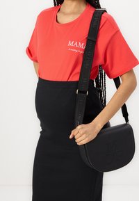 Žena v červeném tričku s nápisem "MAMA na tebe čeká", černé žebrované sukni a černé crossbody tašce s detaily pásku "MOY Polo".