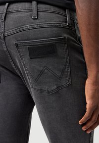 Bližnji posnetek osebe, ki nosi temno sive jeans hlače Wrangler, prikazuje zadnji žep z našitim "W" in blagovno znamko, desna roka sproščena ob strani.