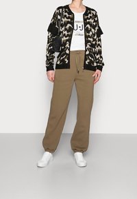 Cárdigan de camuflaje tejido con ribete negro, camiseta gráfica blanca y pantalones deportivos marrones de estilo relajado. Combinado con zapatillas blancas.