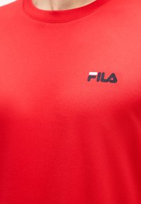 Gros plan sur un maillot de sport rouge texturé avec un col rond et un petit logo FILA sur la poitrine.