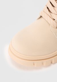 Botte beige avec semelle en caoutchouc épaisse ton sur ton et lacets tressés assortis, présentant des coutures détaillées et une zone de pied arrondie.