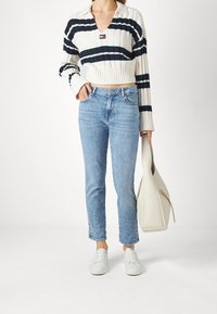 Suéter blanco de punto con rayas azul marino, diseño recortado, escote en V, combinado con jeans azul claro y zapatillas blancas. Sosteniendo un bolso de lona beige.