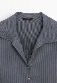 Chemise bleu marine avec fines rayures verticales blanches, col ouvert et deux boutons métalliques visibles sur la patte de boutonnage avant.