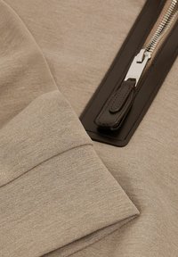 Beige collegepaita, jossa on sileä pinta, tummanruskea vetoketju nahkaiseen vetotankoon ja erottuvaa kangaspintaa.