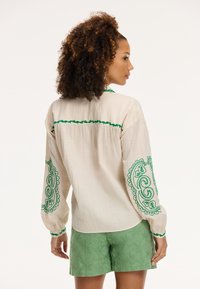Blusa bianca dal texture con accenti ricamati verdi sulle maniche e sul colletto; abbinata a pantaloni corti leggeri con motivo floreale verde.
