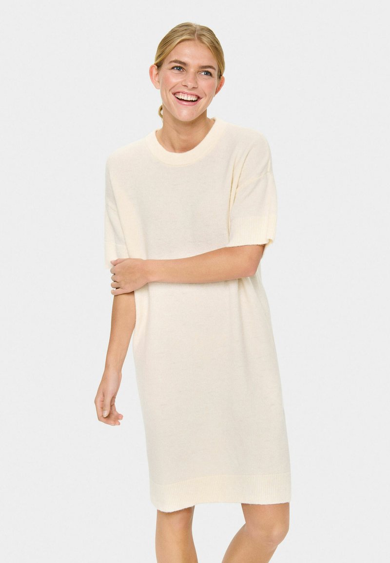 Jeune femme souriante, portant une robe en tricot crème à manches courtes et amples, se tenant devant un fond blanc uni.