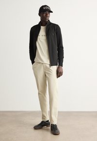 Jachetă neagră cu fermoar și accente aurii, tricou bej cu grafică, pantaloni bej deschis și adidași negri. Pălăria prezintă un logo reliefat.