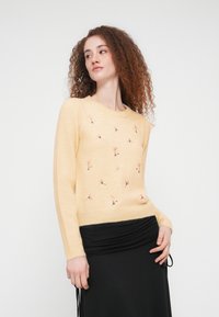 Pull tricoté jaune pâle avec des broderies florales dispersées en rose et vert. Manches longues et poignets côtelés avec un col rond.