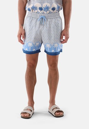 Mann trägt blau-weiß gemusterte Shorts mit Palmenmotiv, steht in beigen Sandalen vor einfarbigem Hintergrund.