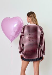 Bordo ir rožinė juostuota ilgomis rankovėmis marškiniai su tekstu ant nugaros, derinami su pilka plisuota sijonu ir rožiniu širdies formos balionu.