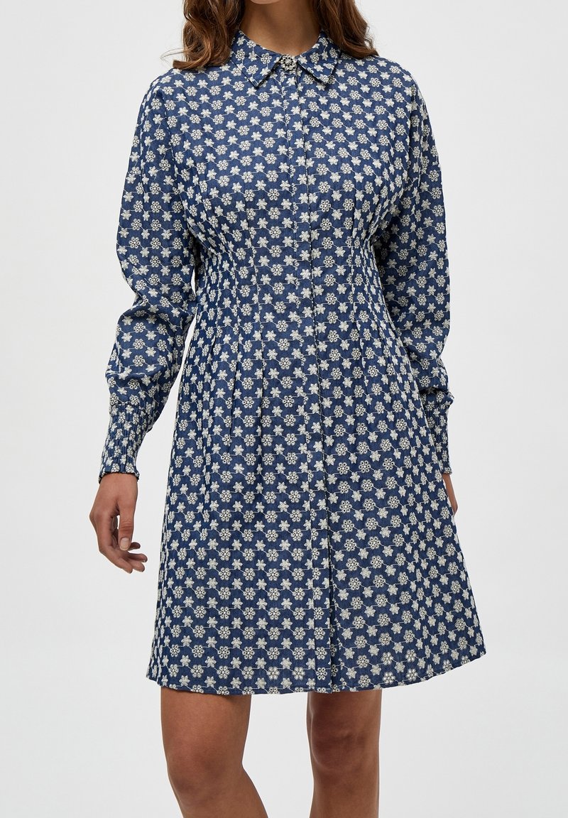 Robe bleu marine avec motif floral blanc, devant à boutons, col, et manches bouffantes longues. Le tissu semble léger avec une texture douce.