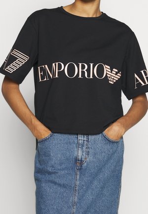 Personne portant un T-shirt noir Emporio Armani avec un logo rose et une jupe en jean bleue, les mains dans les poches, cadrée au niveau du cou et des genoux.