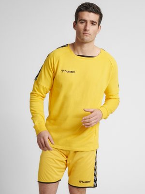 Mand iført gult hummel sportswear med sorte chevronstriber på skuldre og shorts, stående mod en ensfarvet grå baggrund.