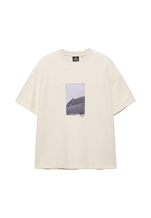 Maglietta oversize color crema con maniche corte e una foto in bianco e nero di una casa sulla collina stampata al centro davanti.