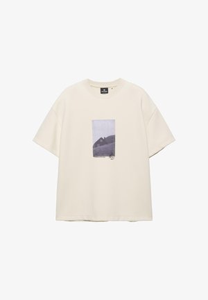 Krém színű, túlméretezett rövid ujjú póló középen, elöl egy fekete-fehér házfotóval, amely egy dombon áll.