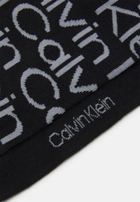 Berretto in maglia nera con design del testo del logo grigio chiaro e branding "Calvin Klein" sul bordo inferiore. Il materiale sembra morbido e elasticizzato.