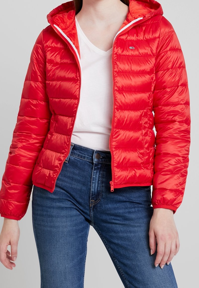 Femme portant une veste matelassée à capuche rouge vif par-dessus un t-shirt blanc à col en V et un jean bleu, debout devant un fond gris uni.