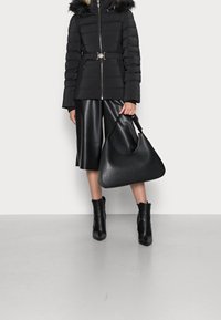 Doudoune noire avec capuche à fourrure, taille ceinturée ; associée à des culottes en cuir noir et des bottines ; un grand sac en cuir est tenu.