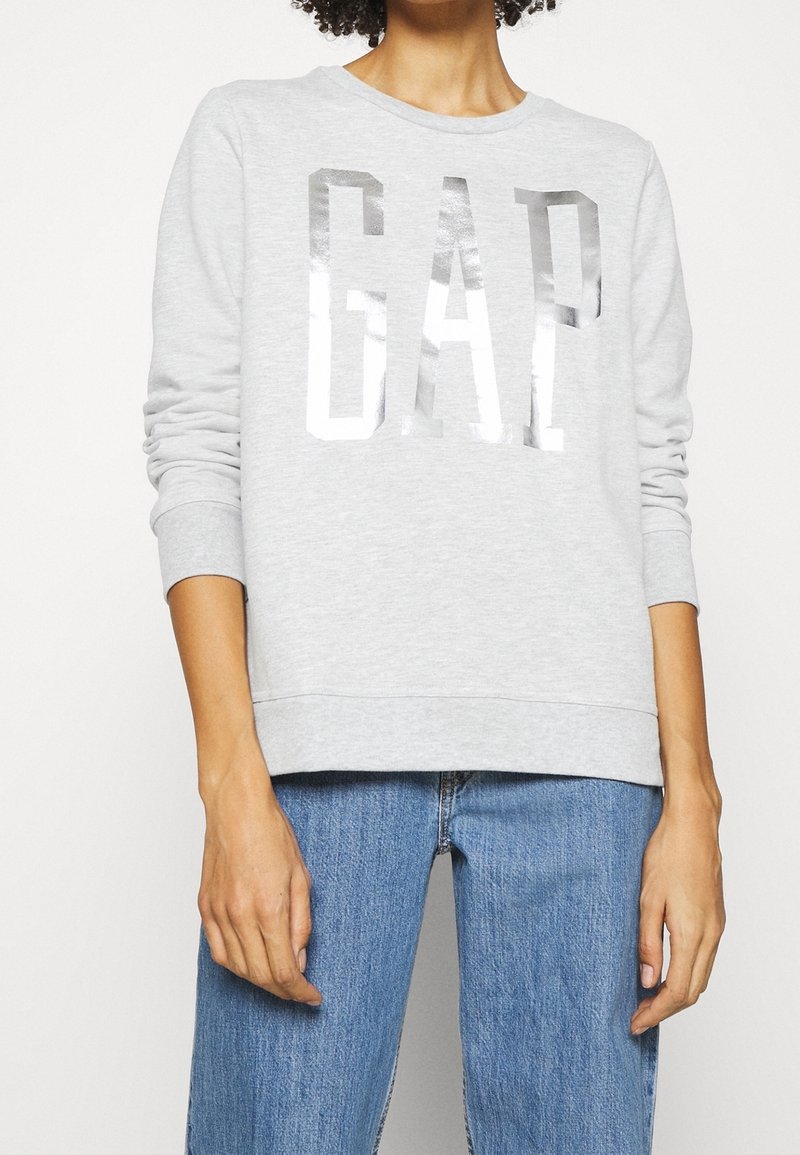 Sudadera gris con cuello redondo, que presenta grandes letras plateadas "GAP". Confeccionada en una tela suave con puños y dobladillo acanalados. Ajuste recto.