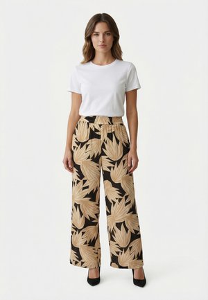 Femme debout portant une chemise blanche à manches courtes et un pantalon large à motifs de feuilles beige, ainsi que des talons hauts noirs, sur un fond uni.