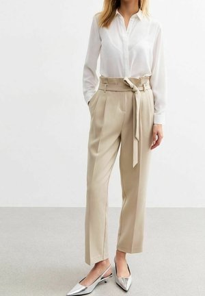 Kvinde iført beige bukser med høj talje og bindebånd, hvid skjorte med knapper og sølvfarvede spidse slingback-sko, stående med hænderne i lommerne.