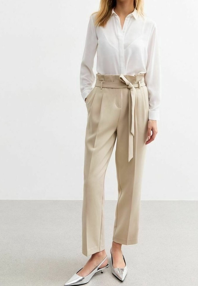 Femme portant un pantalon beige taille haute avec ceinture nouée, chemise blanche boutonnée, et chaussures slingback argentées à bout pointu, debout les mains dans les poches.