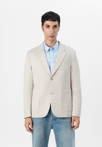 Beiger Strickblazer mit strukturiertem Design, zwei vorderen Taschen und braunen Knöpfen, getragen über einem hellblauen Hemd und Jeans.