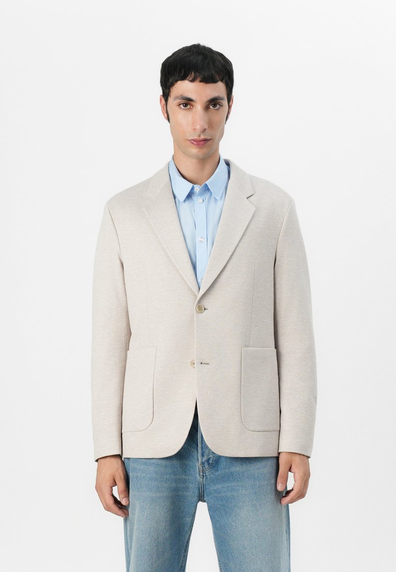 Beiger Strickblazer mit strukturiertem Design, zwei vorderen Taschen und braunen Knöpfen, getragen über einem hellblauen Hemd und Jeans.