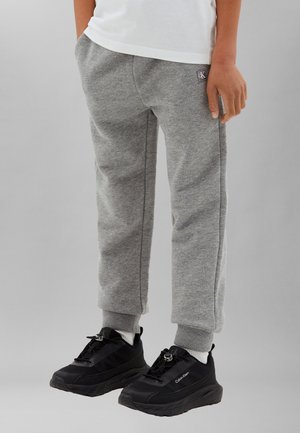 Dziecko w szarych spodniach dresowych Calvin Klein i czarnych butach sportowych Calvin Klein stoi z jedną ręką w kieszeni na jednolitym tle.