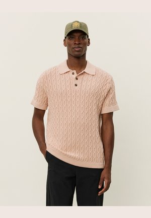 Lys rosa strikket polo skjorte med et teksturert mønster, korte ermer, og en krage med tre brune knapper. Båret med svarte bukser.