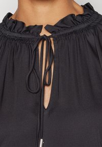 Czarna bluzka z falbaniastym wykończeniem dekoltu i zapięciem na troczki. Gładki materiał, otwór typu keyhole oraz marszczenia wokół kołnierza.