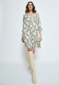 Weißes Kleid mit grün-braunem Blumenmuster, V-Ausschnitt, Dreiviertelärmel und rüschengesäumtem Saum. Kombiniert mit knielangen beigen Stiefeln.
