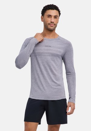 Mann trägt ein hellgraues langärmeliges Sporthemd und schwarze Shorts, posiert mit einer Hand in der Nähe des Kragens.